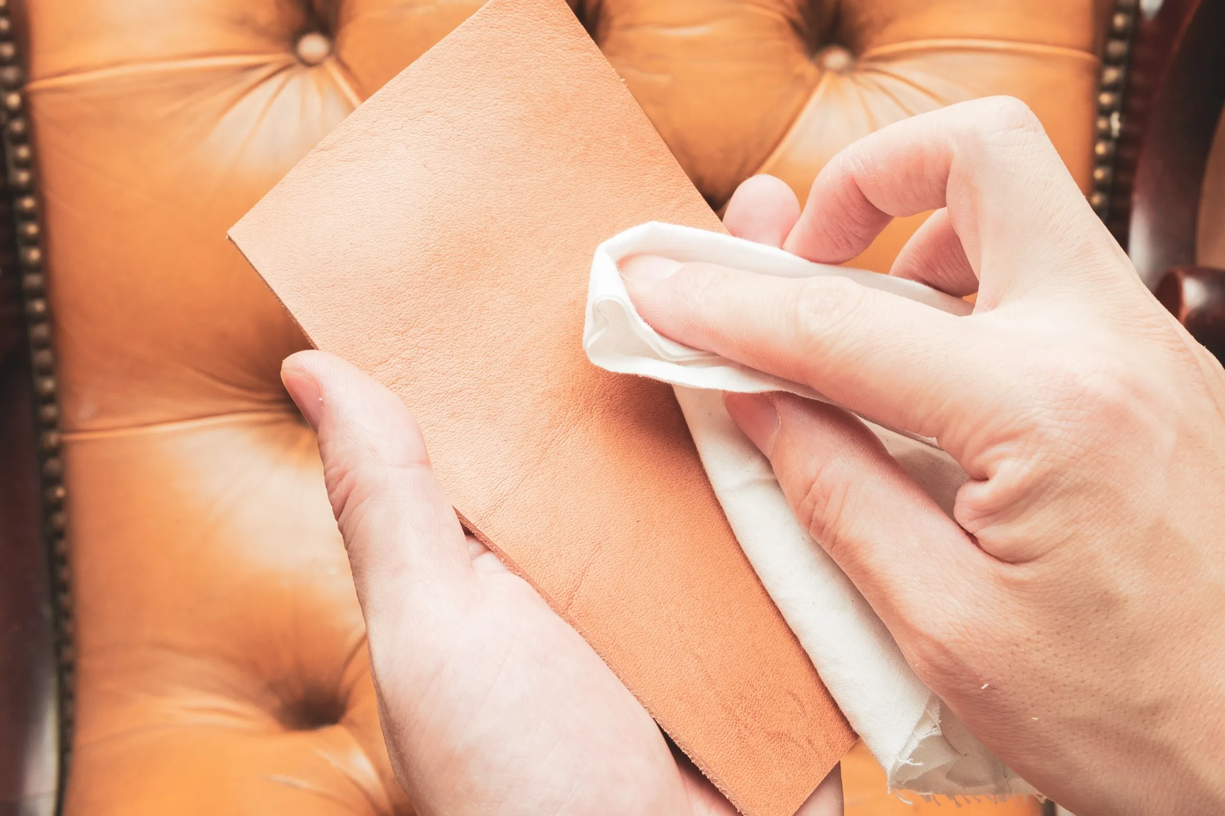 Hands wiping a leather swatch with a soft cloth over a tufted leather chair