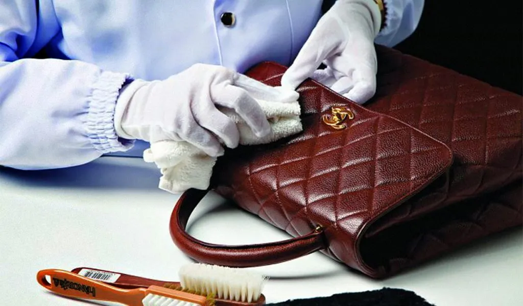 Gloved hands conditioning a quilted burgundy leather handbag